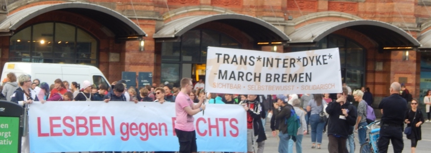 Menschen versammeln sich vor dem Hauptbahnhof. Das große "LESBEN gegen RECHTS"-Banner ist gut zu sehen. Vorne das weiße Banner "Trans*Inter*Dyke*-March Bremen"